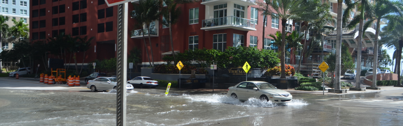 slide 2 - Downtown Miami scene shows a car moving through a flooded intersection on a sunny day; a stop sign looms in the foreground, buildings and palm trees in the distance, and the water, though fairly shallow, shows small wavelets.