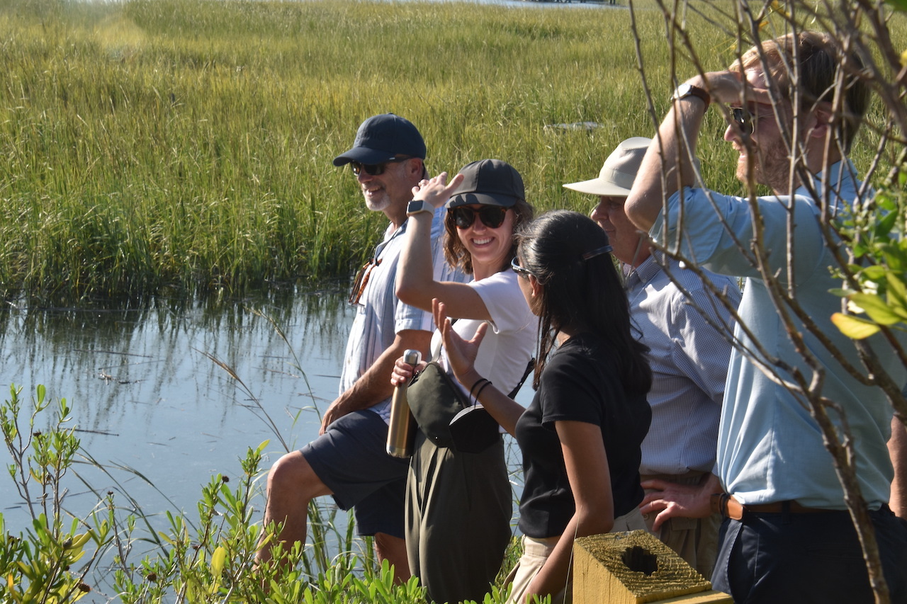 A cluster of people who attended a NASA Sea Level Change Team meeting in September stands among wetland reeds and shrubs, next to a water channel, in bright sunlight on Tybee Island. The tour group, some shielding their eyes, look to the left toward a water management structure (off screen).