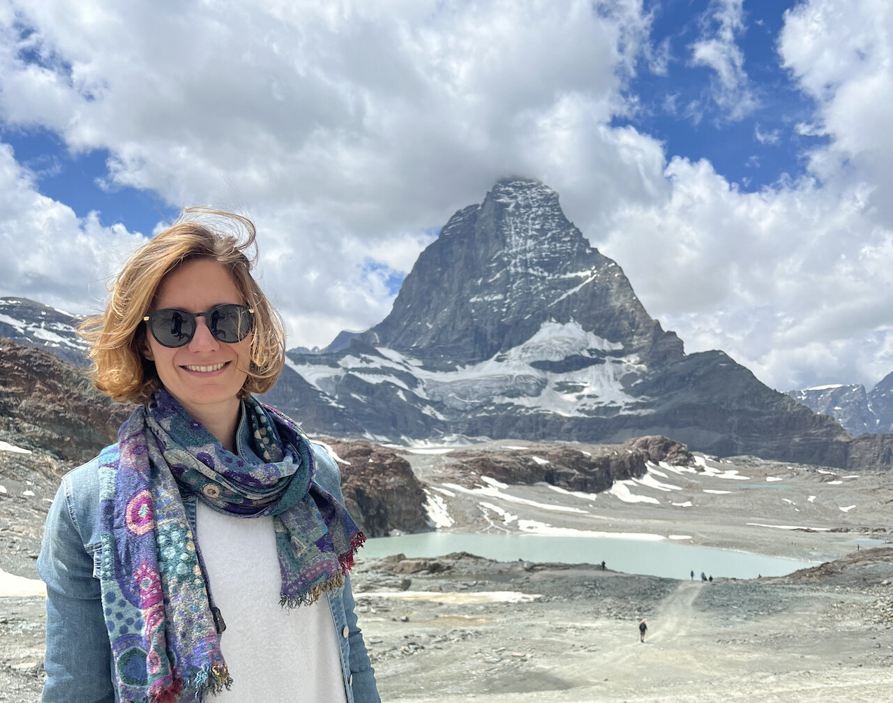 Researcher Manuela Girotto, wearing sunglasses and scarf, stands to the left of an icy mountain above a lake of melt-water.